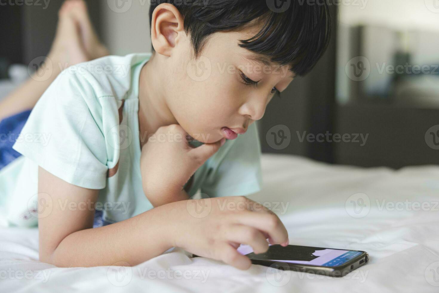 Close up portrait of Asian boy with black bangs, black eyes with a smiling face wearing a light green shirt lying on the bed in his house playing with his smart phone. Education concept. photo