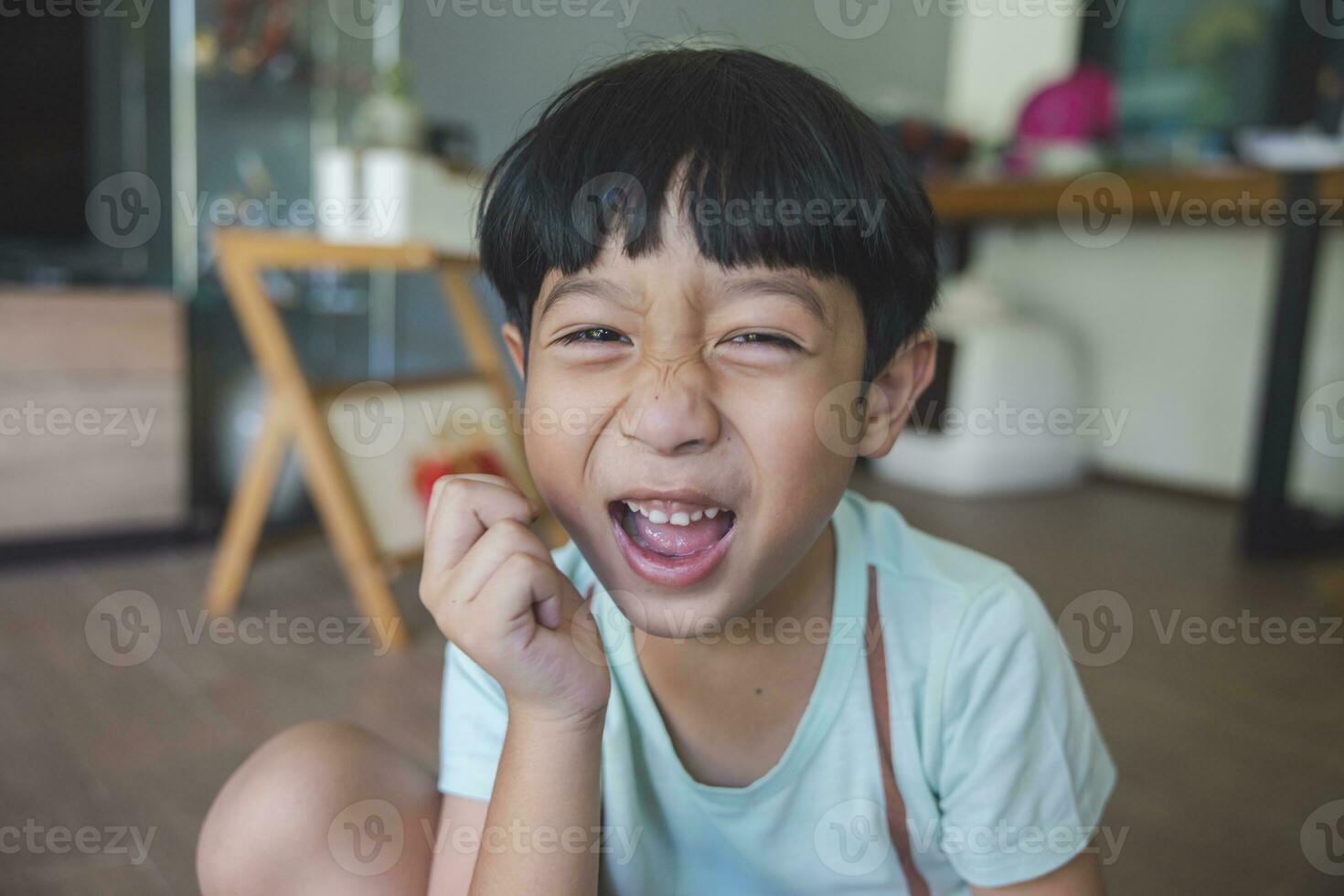 Close up portrait of Asian boy with black bangs, black eyes with a smiling face wearing a light green shirt look at camera and sitting on the floor of his house and hand holding pencil to draw. photo