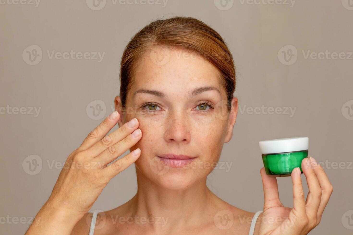 Portrait of caucasian middle aged woman face with freckles showing soft skin by fingers holding cream photo
