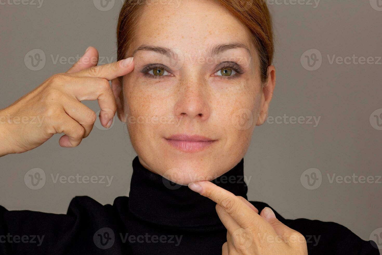 retrato de recortado caucásico sonriente medio Envejecido mujer cara con pecas demostración en párpado y barbilla por índice dedos foto