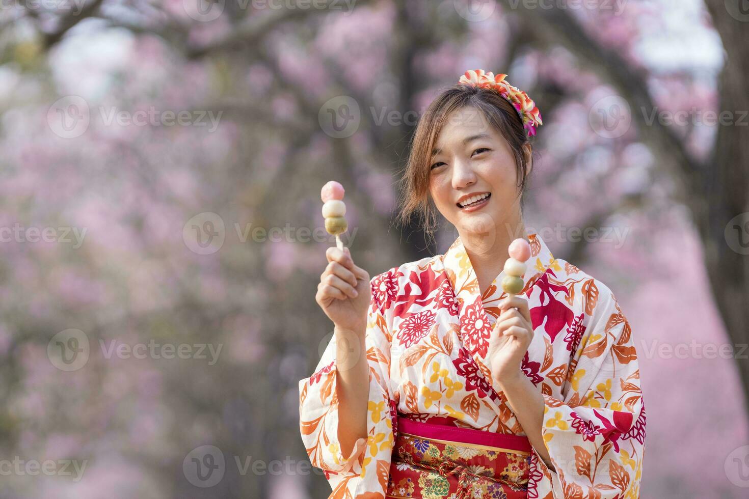 Japanese woman in traditional kimono dress holding sweet hanami dango dessert while walking in the park at cherry blossom tree during spring sakura festival photo