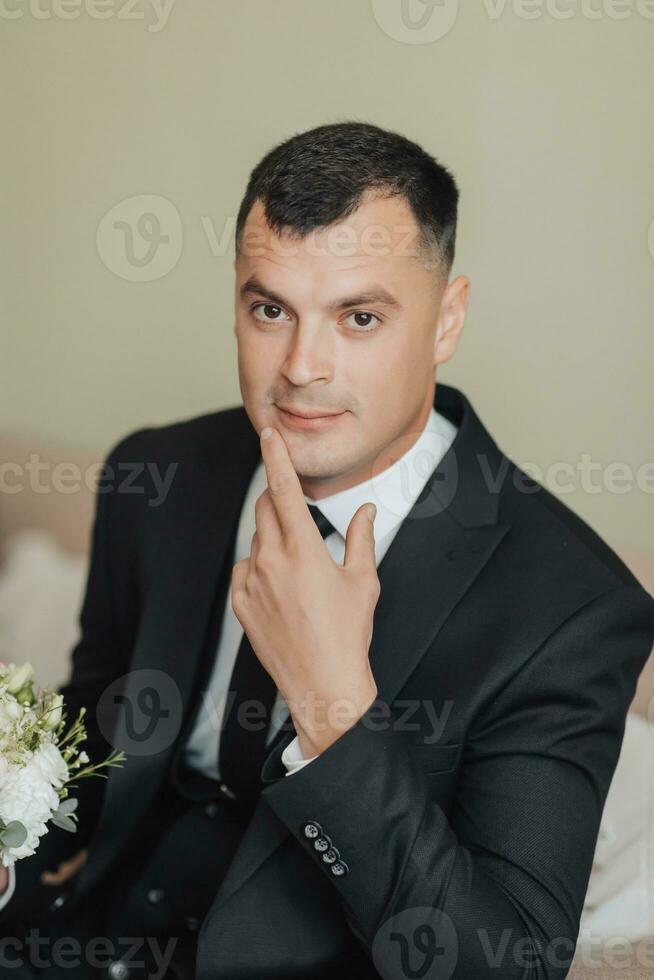 retrato. un hombre en un blanco camisa, negro Corbata y negro clásico traje se sienta en un marrón sofá con un ramo de flores de blanco rosas. un elegante mirar. de los hombres estilo. moda. negocio foto