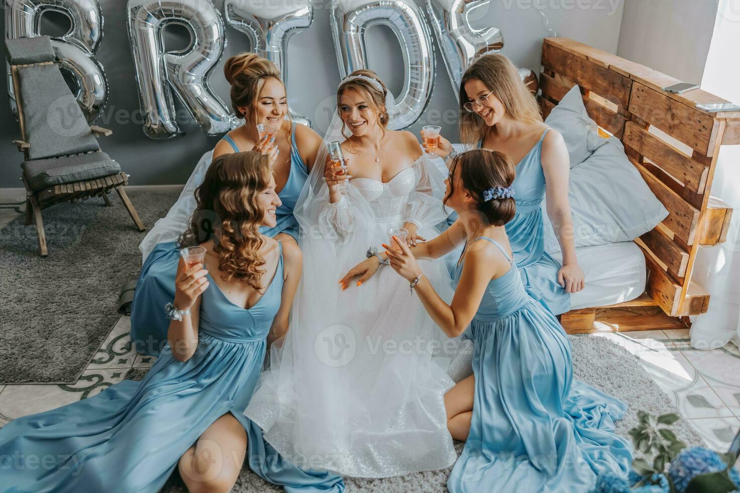 joven damas de honor en azul seda vestidos bebida champán en el de la novia habitación. hermosa mujer celebrando despedida de soltera fiesta sentado en cama y con champán. foto