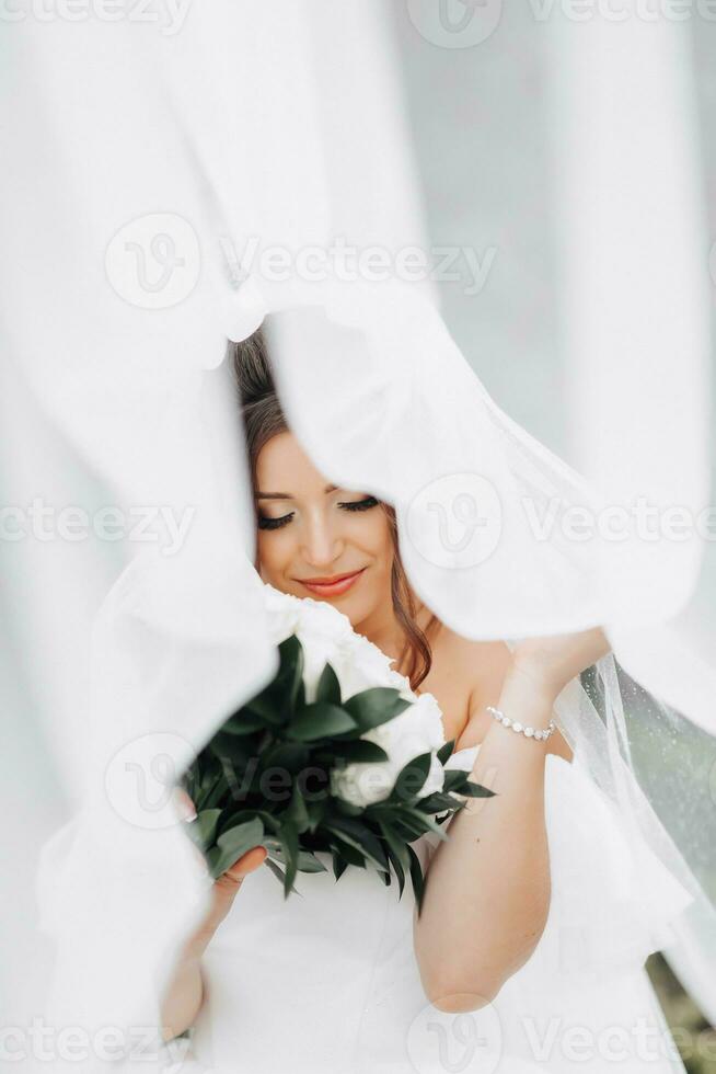 Curly brunette bride in a white dress, covered with a veil, poses for the camera with a bouquet of roses. Portrait of the bride. Beautiful makeup and hair. Wedding in nature photo