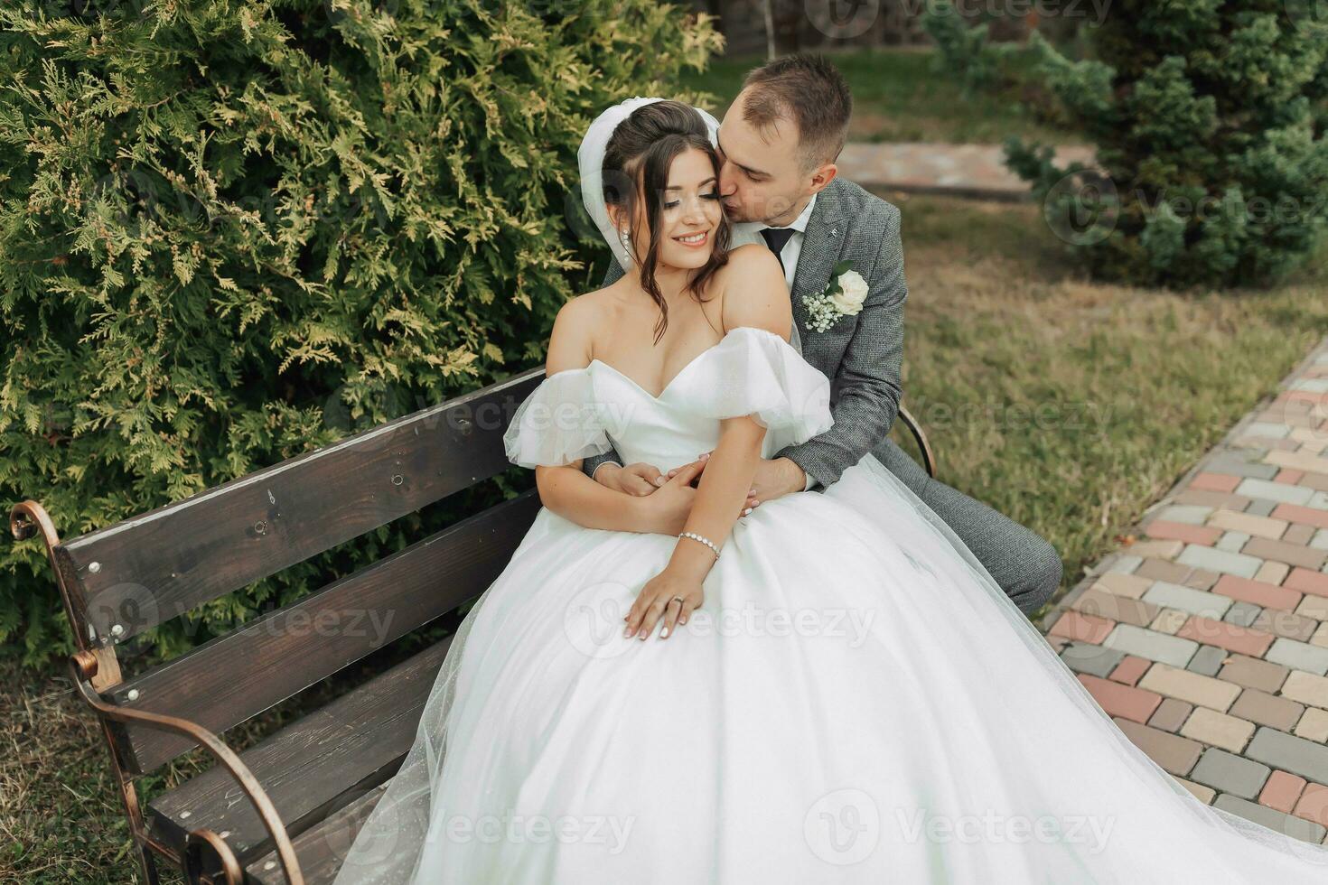 Portrait of the bride and groom in nature. A stylish groom and a brunette bride in a white long dress sit on a bench against a tree background, hug and sincerely smile. curly hair photo