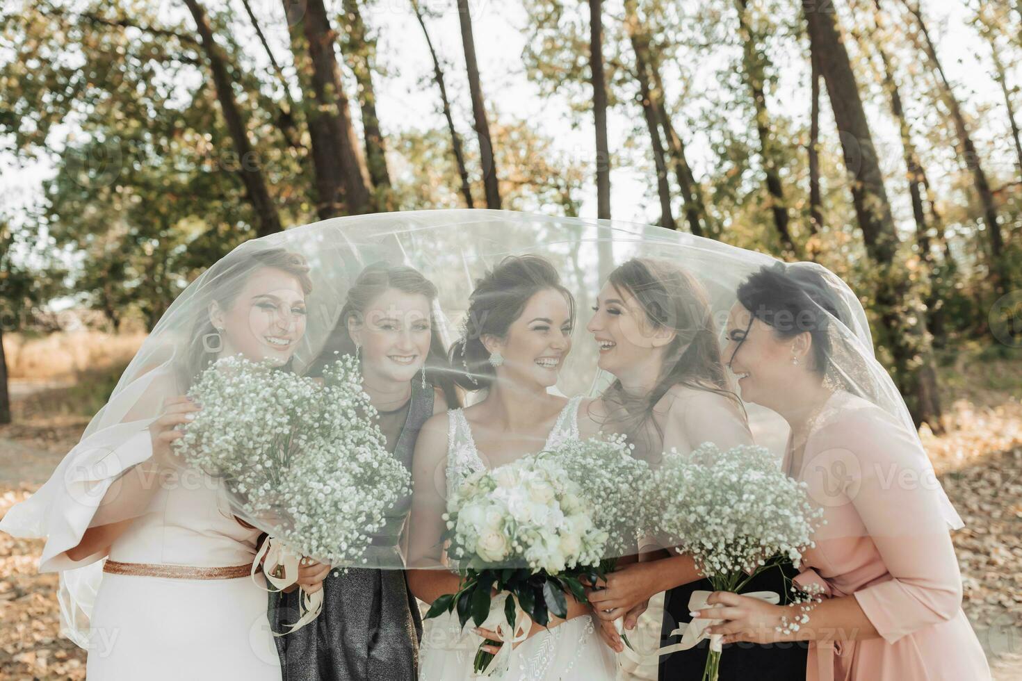 Wedding photo in nature. The bride and her friends are standing in the forest holding their bouquets, smiling, covered by the bride's veil. Wedding portrait. Emotions. Beautiful girls.