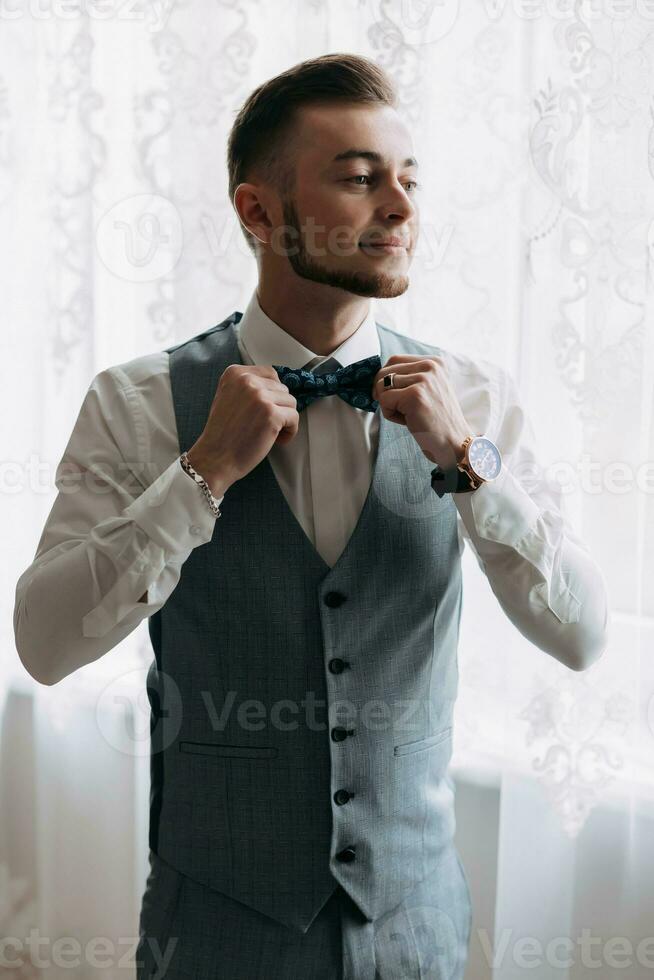 A man in a white shirt and a gray vest poses by the window and looks to the side, adjusts his blue bow tie. Front view. A stylish watch. Men's style. Fashion. Business photo