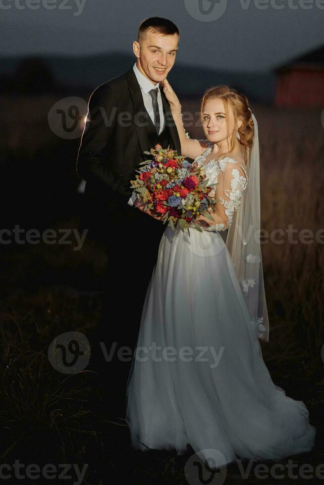 Beautiful bride and groom in evening lights, kissing and hugging holding a bouquet of flowers photo