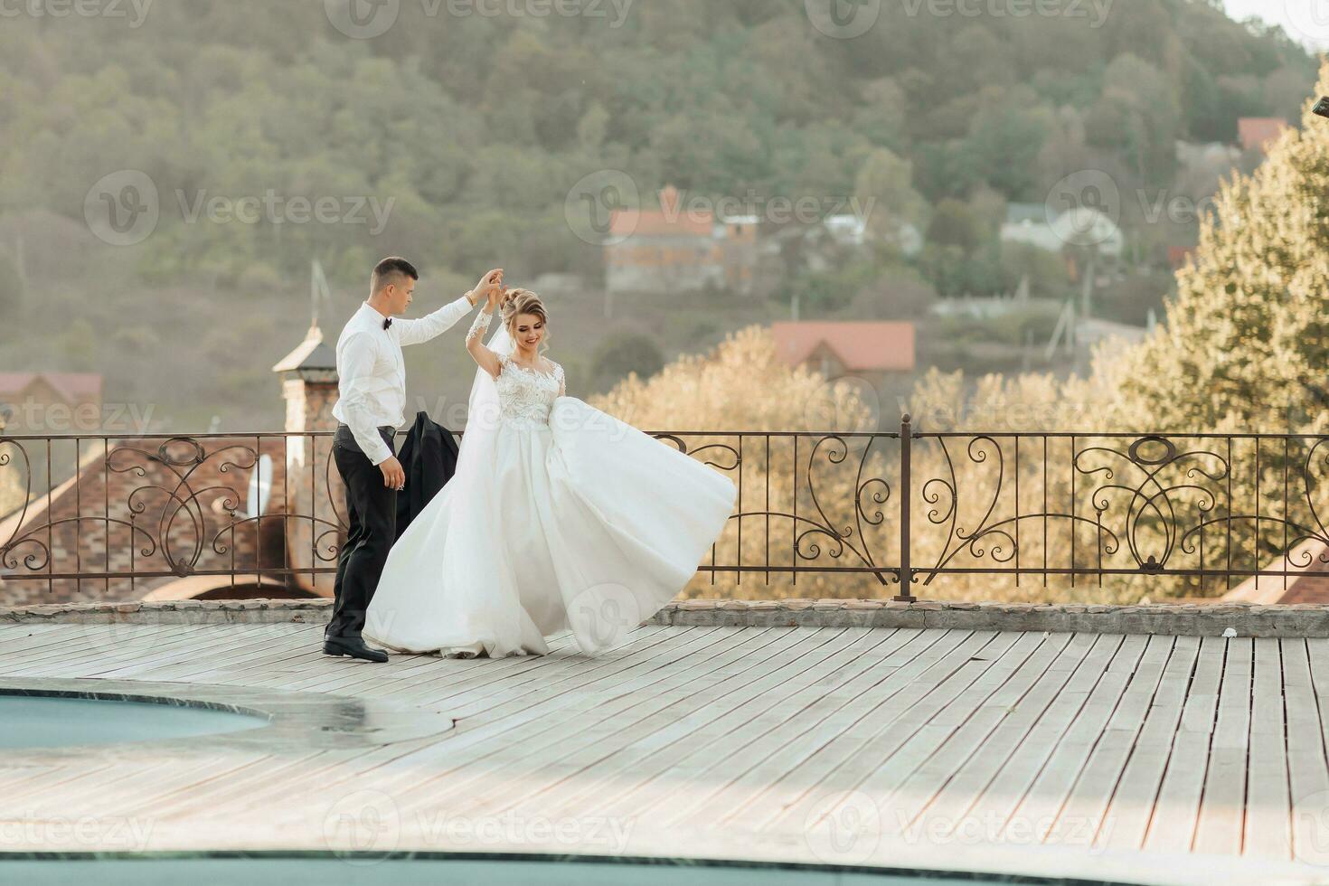 A groom in a black suit and a blonde bride dance a waltz against the background of a mountain and buildings, near a swimming pool. Long dress in the air. Romantic photo in nature. Beautiful hair