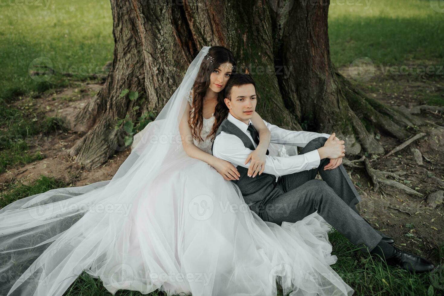 un hermosa novia con largo Rizado pelo en un elegante vestir abrazos el acicalar, sonrisas, mirando dentro el lente debajo un grande árbol. retrato de el novia y novio. primavera boda. natural maquillaje foto