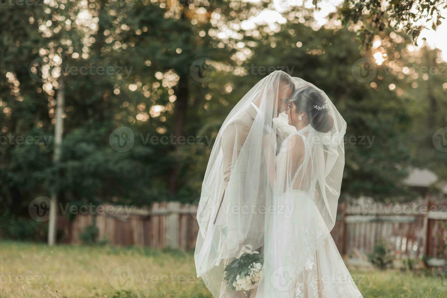 Wedding portrait of the bride and groom. Happy bride and groom gently hug each other under the veil, pose and kiss. Stylish groom. Beautiful young bride photo
