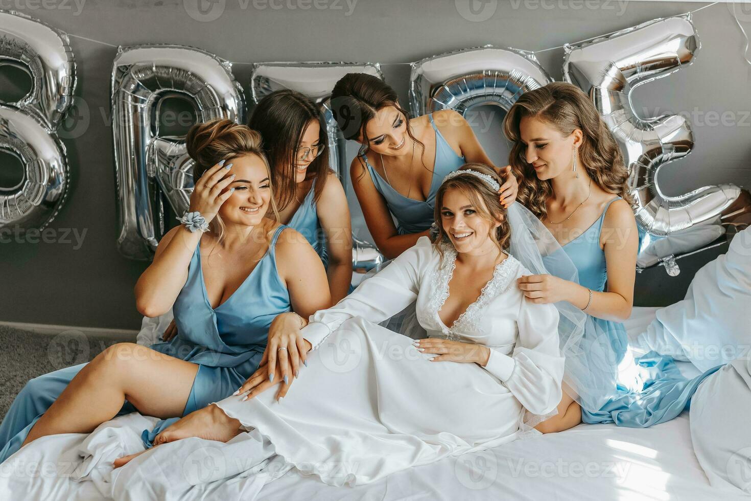 el damas de honor Mira a el sonriente novia. el novia y su alegre amigos son celebrando un despedida de soltera fiesta en el cama en idéntico vestidos. novia y amigos en el habitación foto