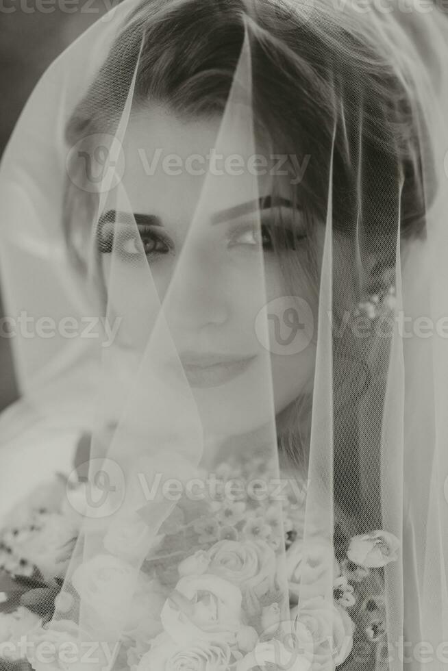 Curly blonde bride in a white dress, covered with a veil, poses for the camera with a bouquet of roses. Portrait of the bride. Beautiful makeup and hair. Wedding in nature. black and white photo