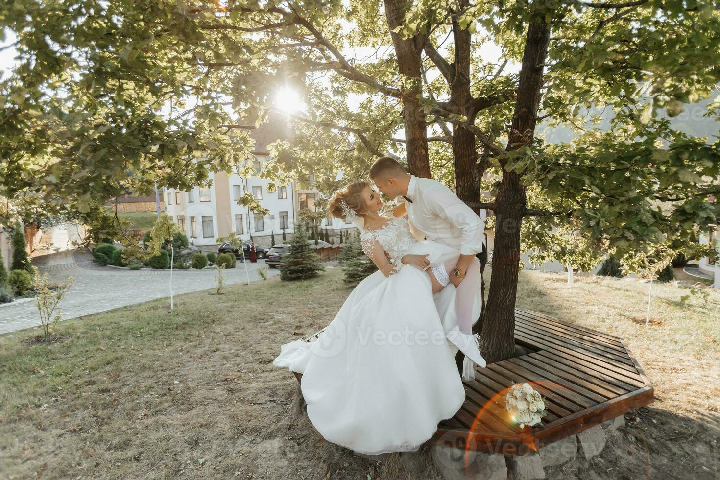 Wedding portrait. The groom in a black suit and the blonde bride are standing, kissing under a tree. The groom passionately holds the bride's leg. Photo session in nature. Beautiful hair and makeup