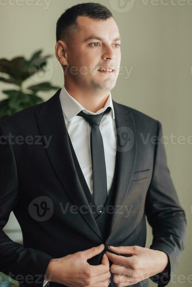 un hombre en un blanco camisa, negro Corbata y negro traje poses por el ventana y mira lejos. arregla su chaqueta. frente vista. un elegante mirar. de los hombres estilo. moda. negocio foto