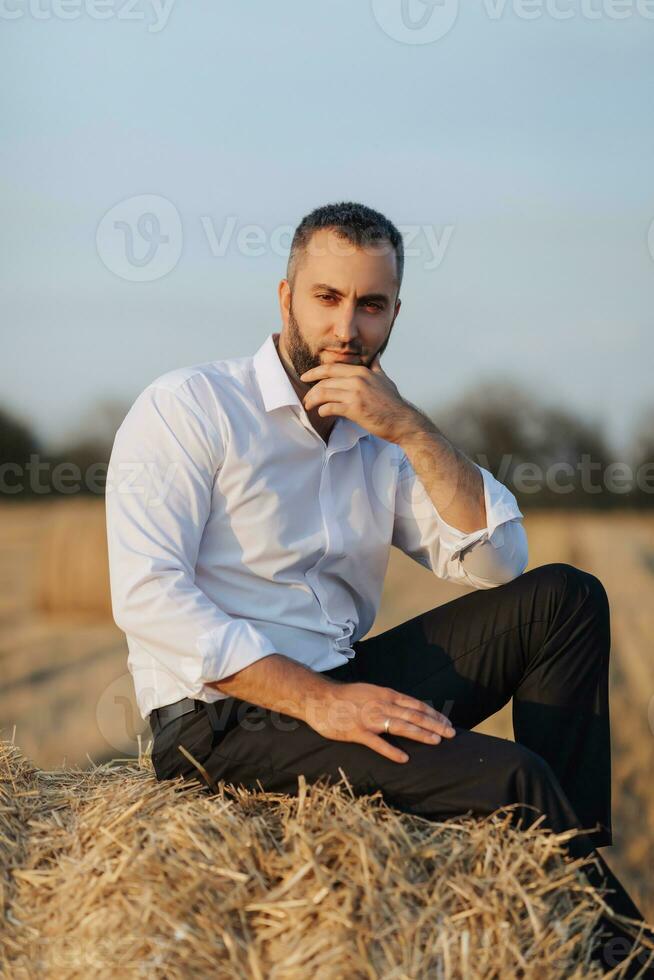 Boda retrato foto. un elegante novio en un blanco camisa se sienta relajado en un heno bala y mira a el cámara. barbado hombre. estilo. foto