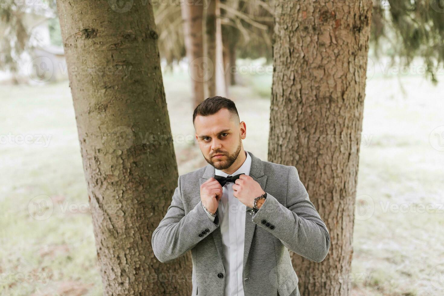 stylish groom, leaning his shoulder against a tree, emphasizes his beard, looks into the camera lens. Portrait photo