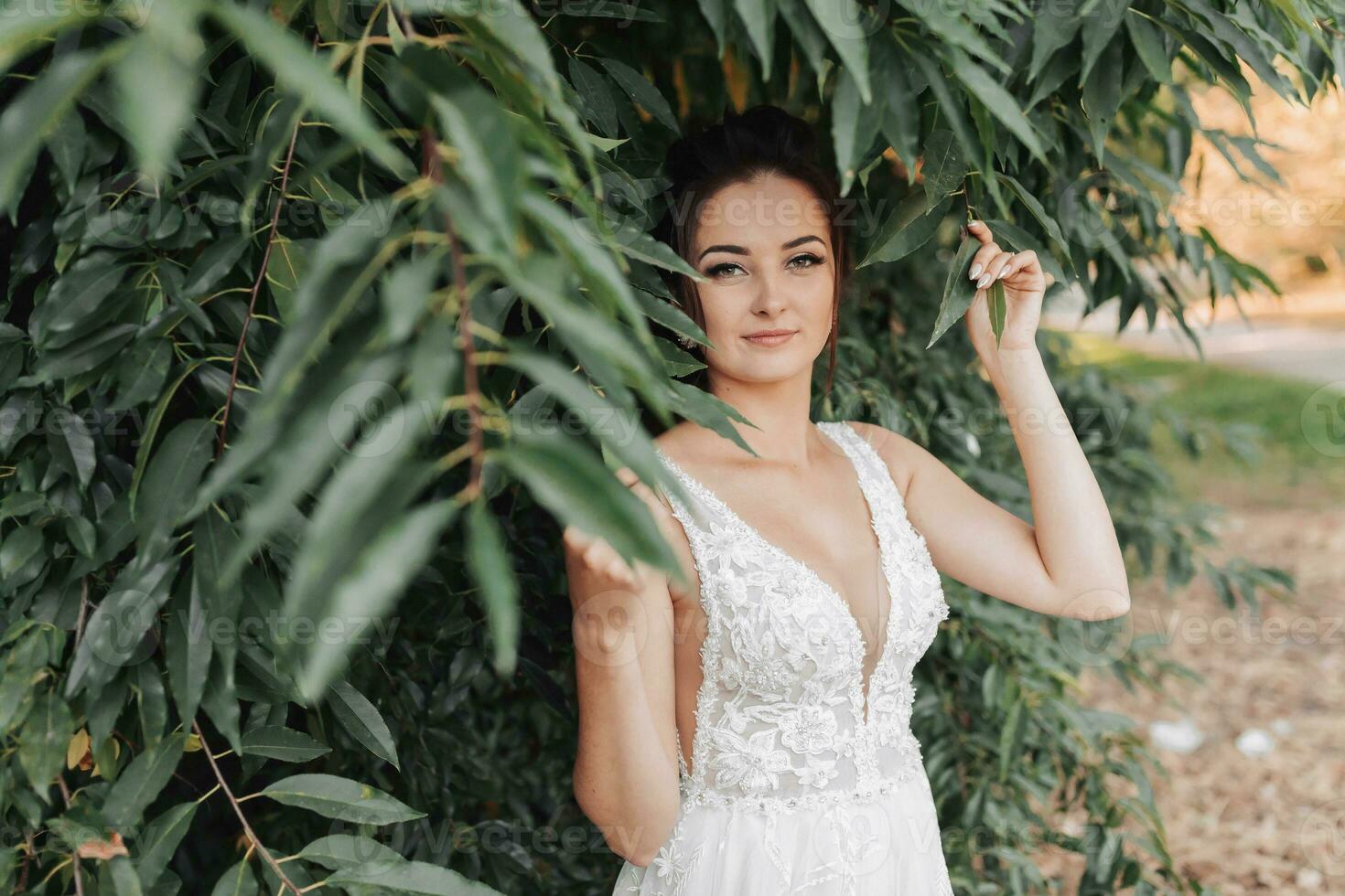 Boda foto en un verde antecedentes. el novia en un blanco vestir soportes cerca verde arbustos, sostiene un hoja y mira dentro el lente. hermosa constituir. retrato de el novia