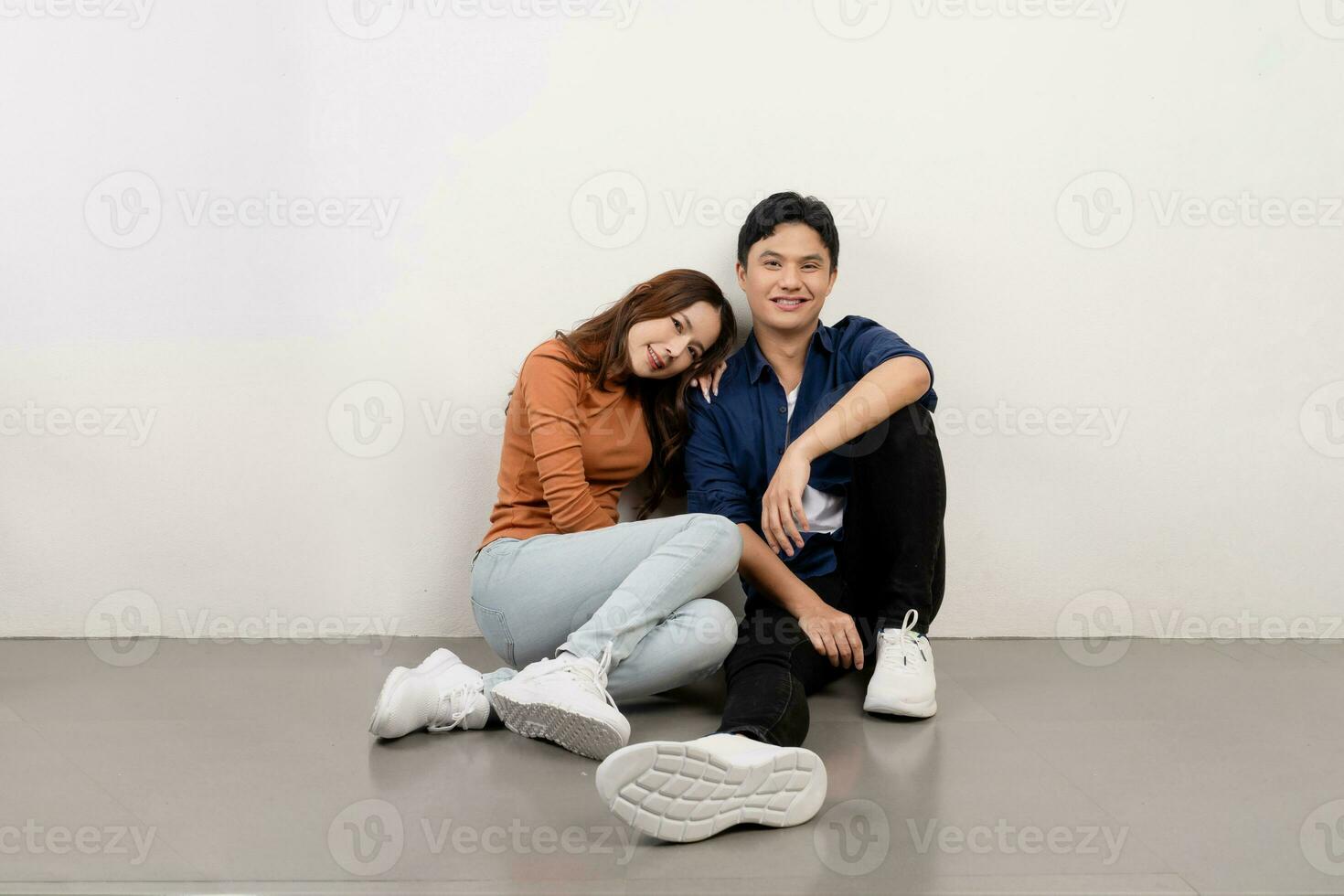 Portrait of cheerful and smiling young Asian couple in casual outfits sitting on floor and looking at camera. photo