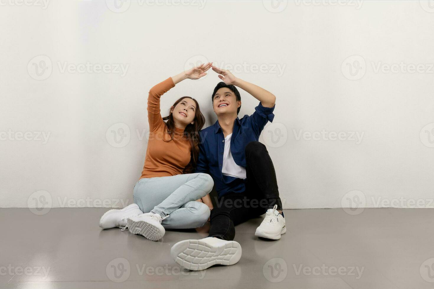 Cheerful young Asian couple in casual outfits sitting on floor and making home roof with their hands, relocation concept photo