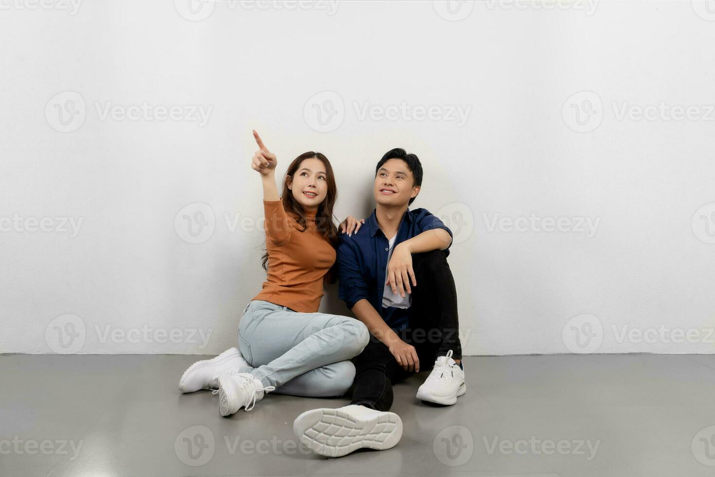 Portrait of cheerful young Asian couple in casual outfits sitting on floor and pointing at copy space photo