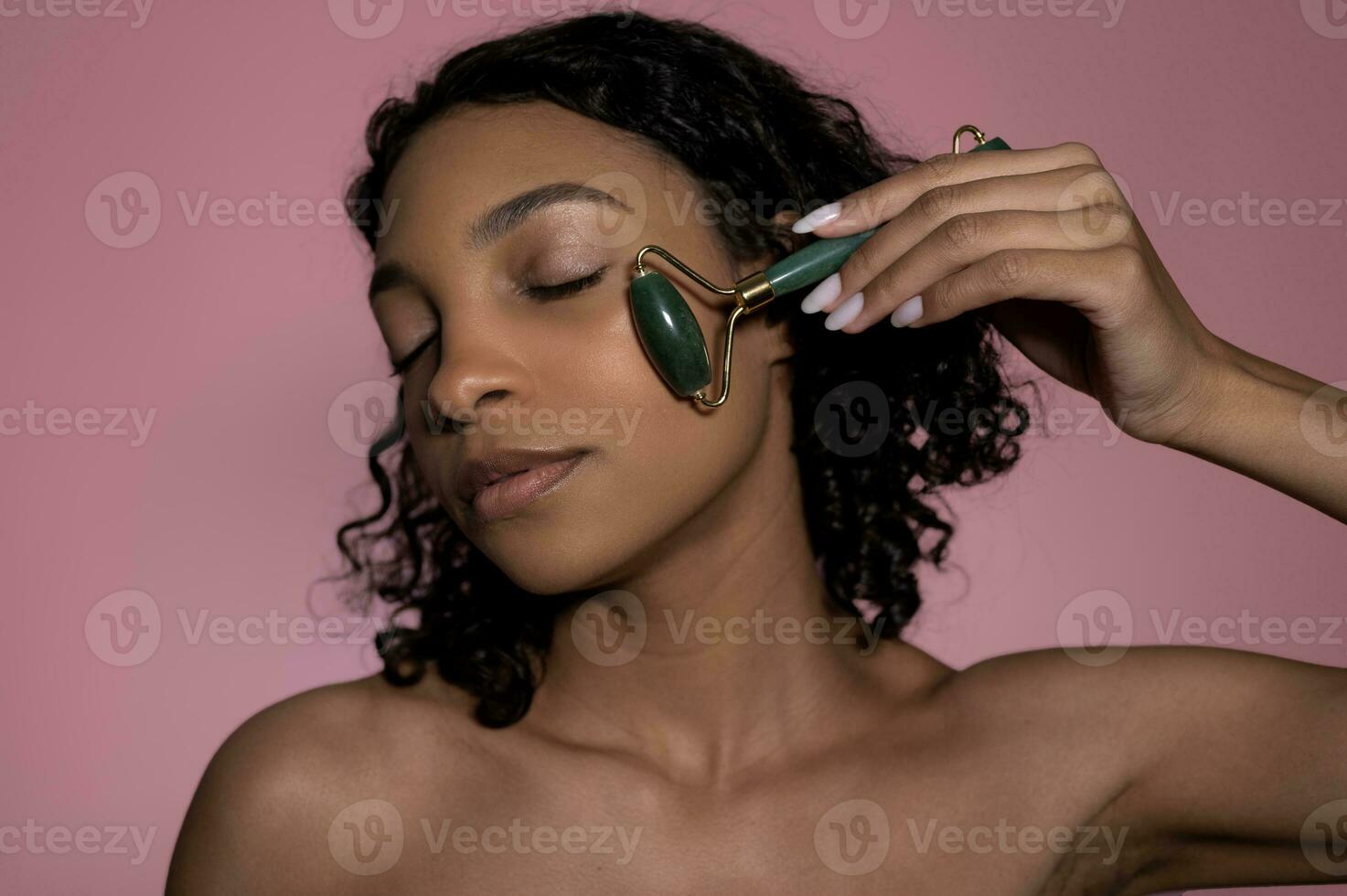 close up view of african american woman with closed eyes using jade roller on face isolated on pink photo