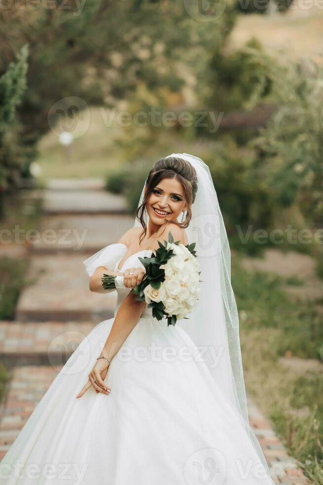Portrait of the bride in nature. A brunette bride in a white long dress, holding a bouquet of white roses, poses, looking into the lens, smiling sincerely. on the background of the tree. Curly hair. photo