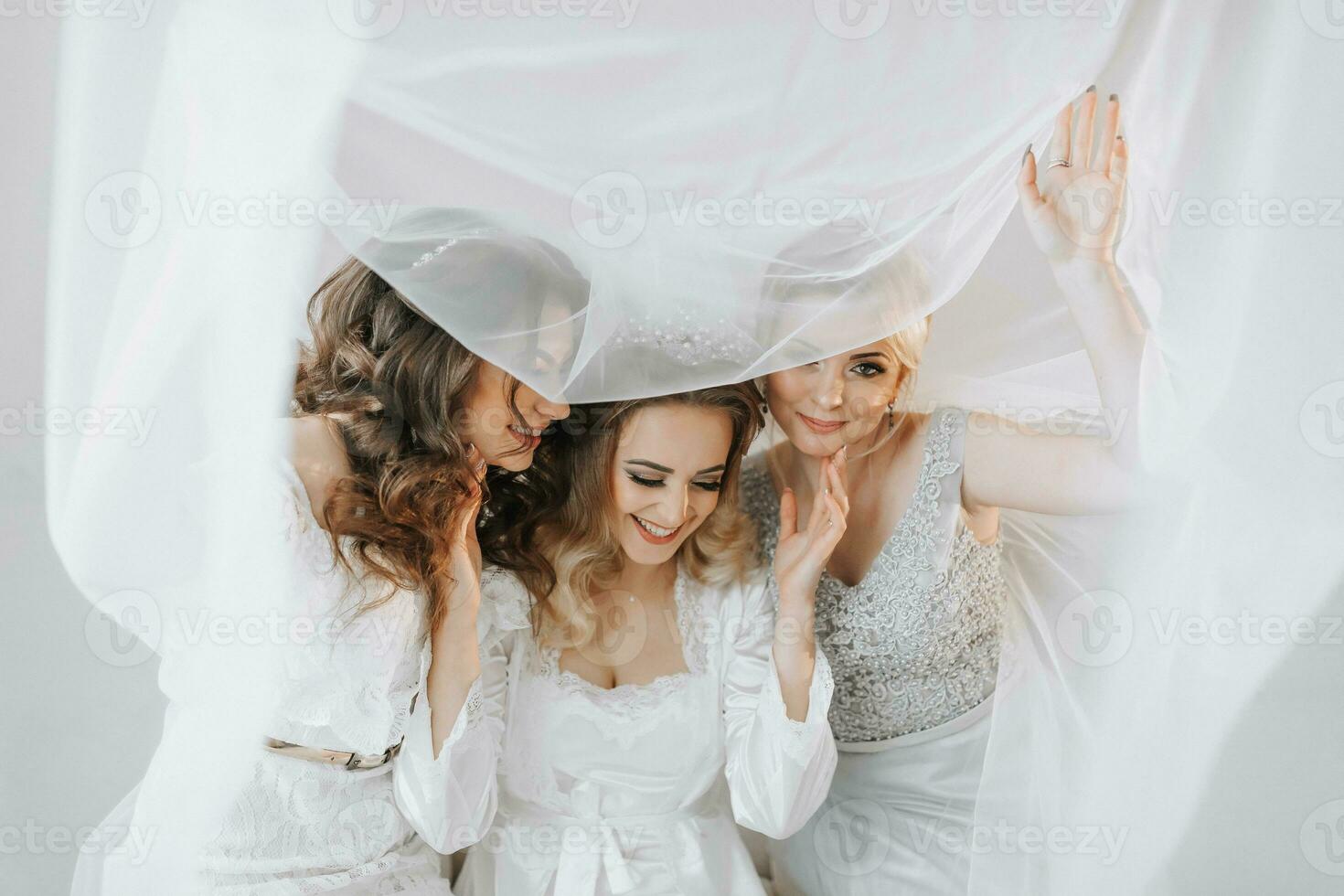 el damas de honor Mira a el sonriente novia. todas en el mismo sofá debajo un velo. el novia y su divertido amigos celebrar el despedida de soltera fiesta en diferente vestidos. novia y amigos en el habitación foto