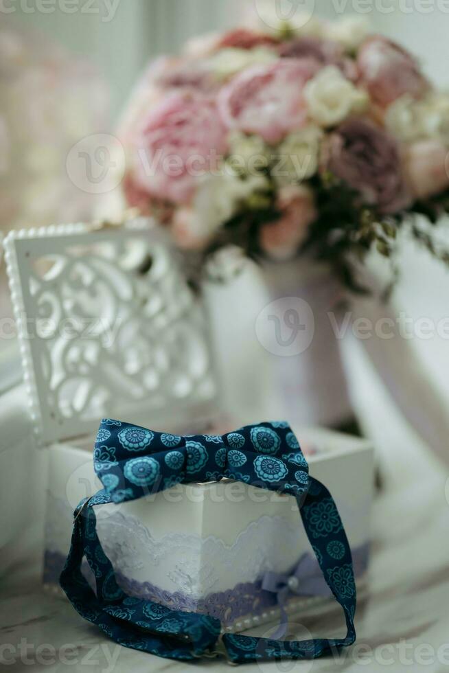 The groom's blue butterfly is standing on a wooden box with wedding rings on the background of a wedding bouquet. Wedding details. photo