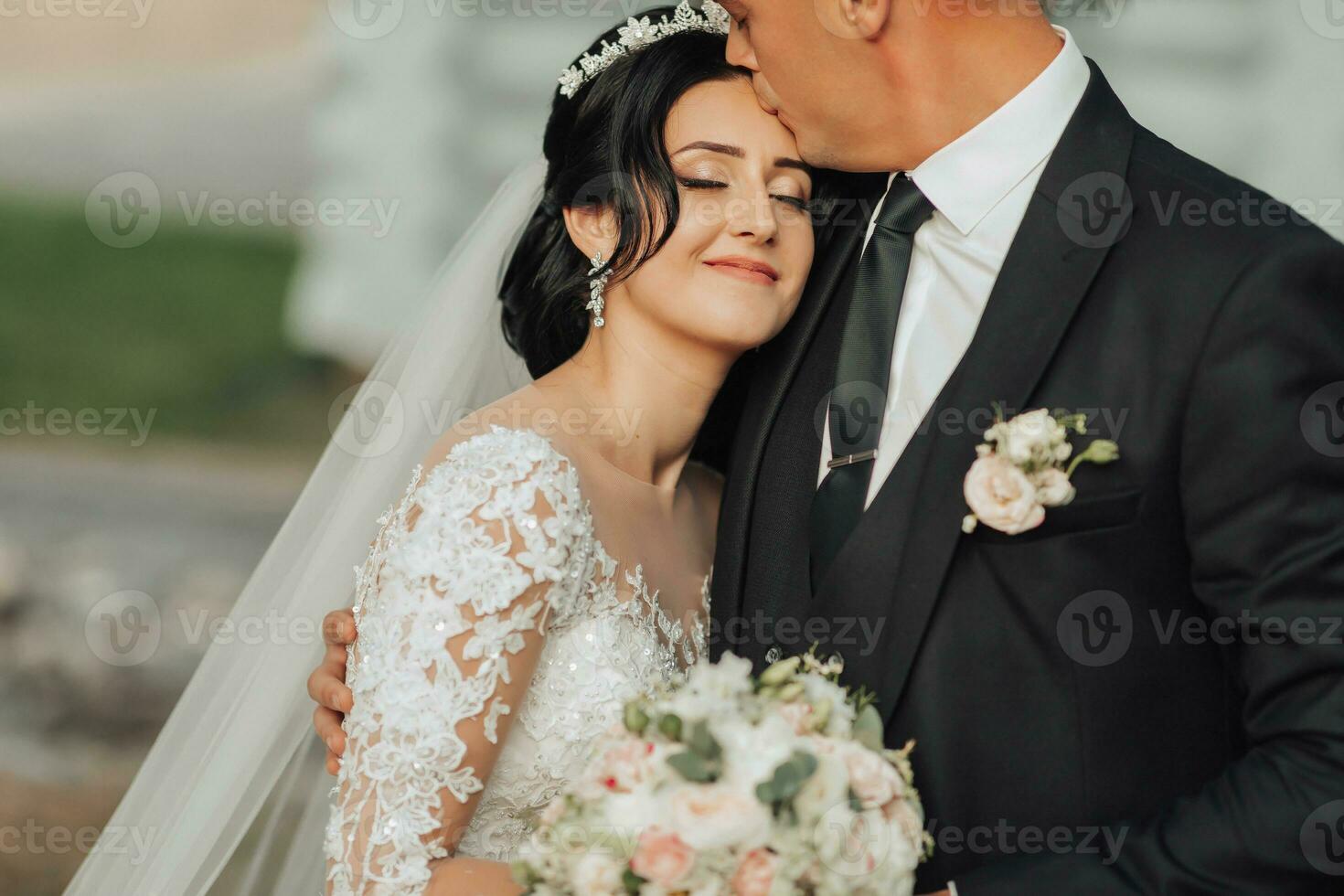 Boda retrato en naturaleza. el morena novia y novio en un blanco largo vestir son en pie y abrazando en contra el antecedentes de conífero arboles y un blanco cabaña. elegante novio. foto