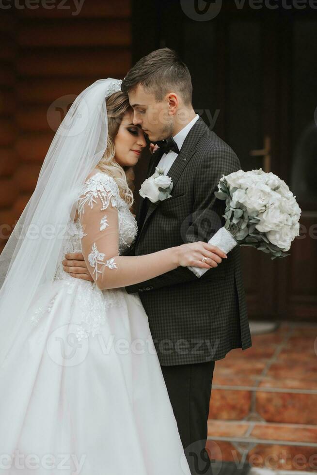 hermoso novio y encantador novia juntos cerca moderno de madera casas en el parque. retrato. magnífico vestir con un largo tren. elegante joven novio foto