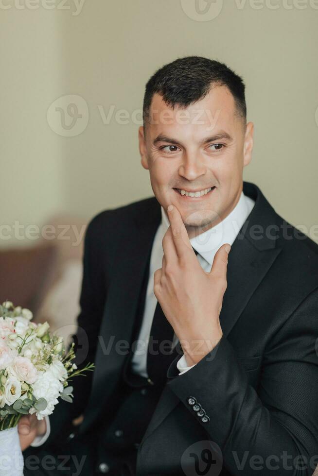 retrato. un hombre en un blanco camisa, negro Corbata y negro clásico traje se sienta en un marrón sofá con un ramo de flores de blanco rosas. un elegante mirar. de los hombres estilo. moda. negocio foto