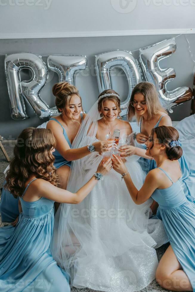 joven damas de honor en azul seda vestidos bebida champán en el de la novia habitación. hermosa mujer celebrando despedida de soltera fiesta sentado en cama y con champán. foto