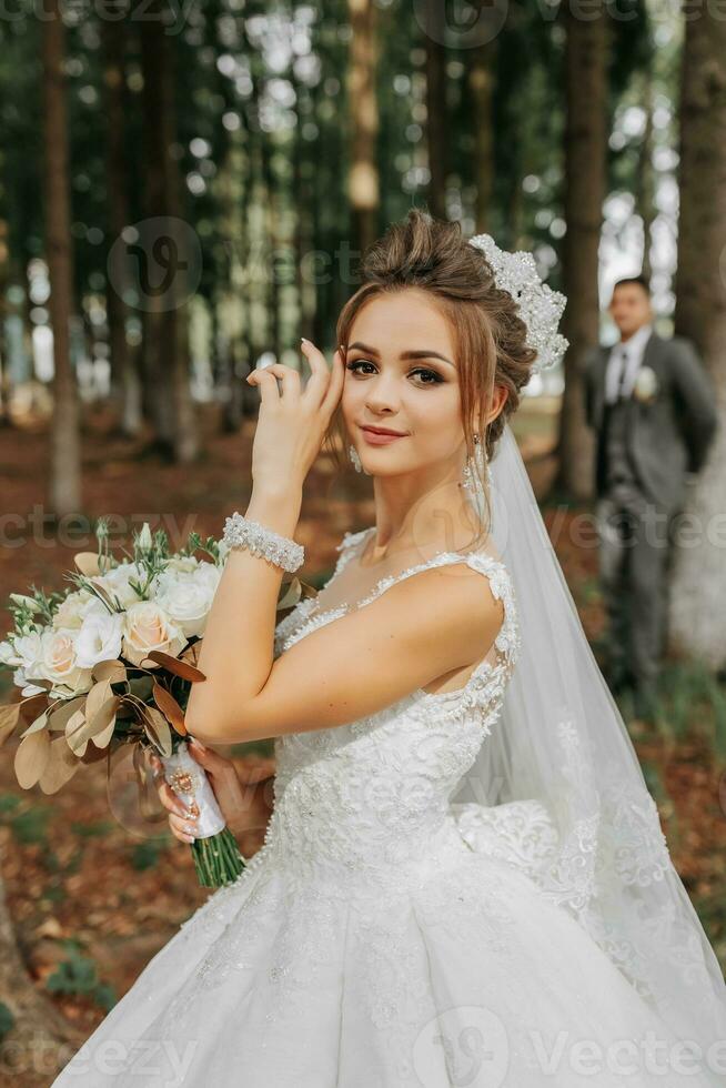 A beautiful young woman in a wedding dress between tall trees in the forest with a royal hairstyle and a chic tiara with a bouquet of flowers in her hands, a wedding in golden color photo