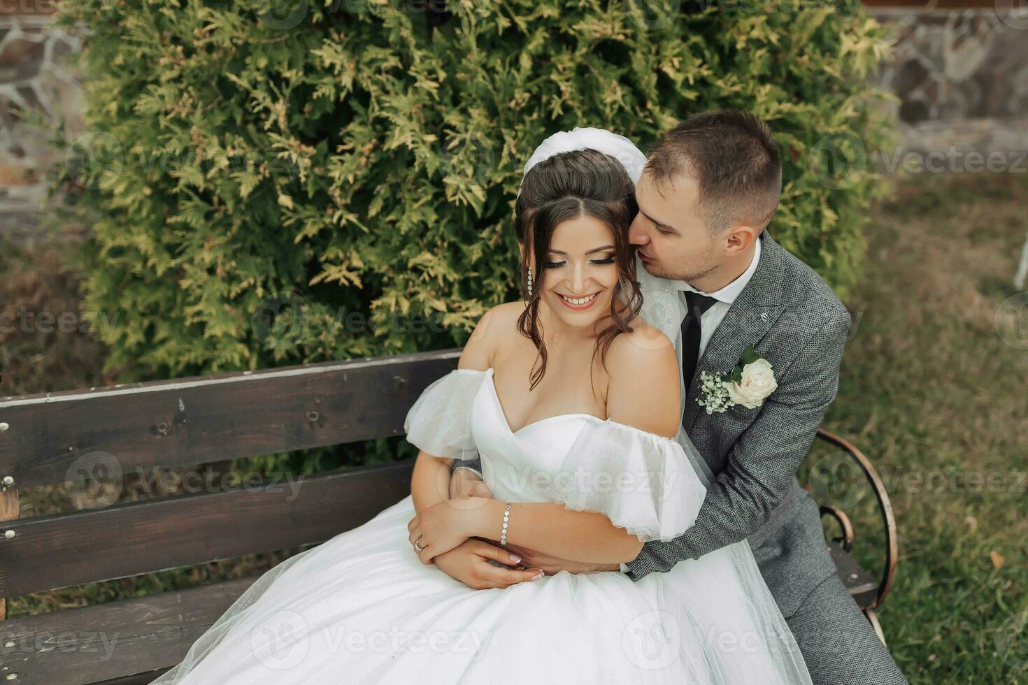 Portrait of the bride and groom in nature. A stylish groom and a brunette bride in a white long dress sit on a bench against a tree background, hug and sincerely smile. curly hair photo