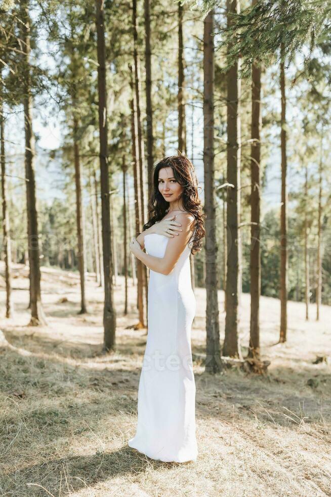 A girl with long black curly hair blowing in the wind is standing in a white silk dress. Standing in the spring forest against the background of large trees photo