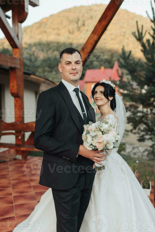 The brunette bride and groom in a white long dress are standing and hugging against the background of coniferous trees and a white hut. Stylish groom. Beautiful hair and makeup photo