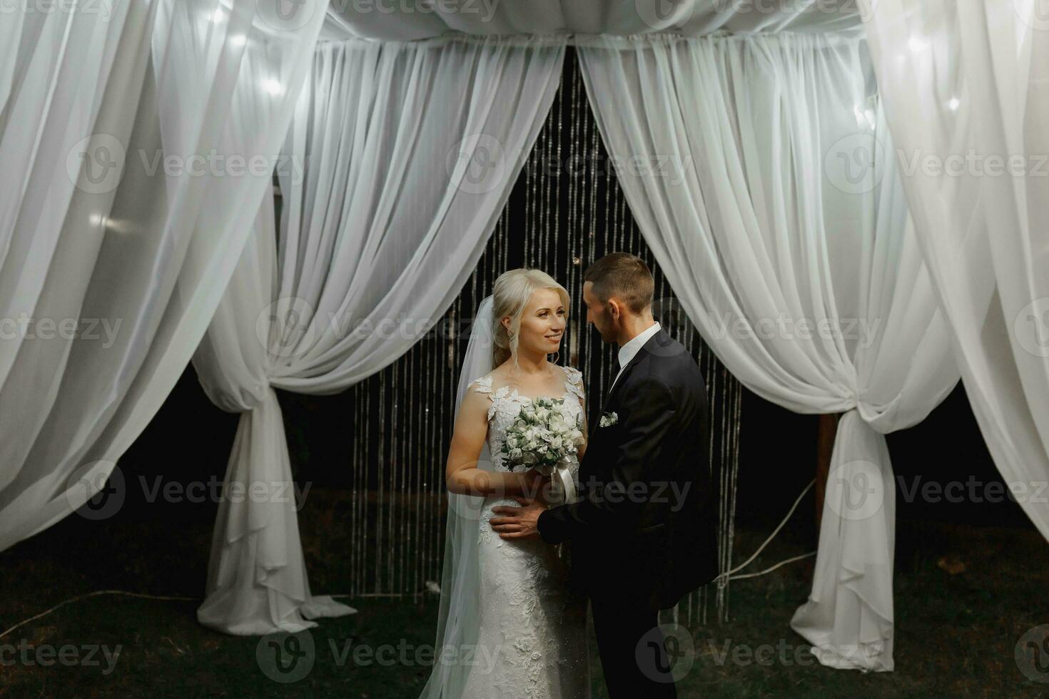 Boda ceremonia en el apuntalar de el lago. pintura ceremonia en el noche. romántico cuadro. el presidio es decorado con blanco flores blanco sillas. el novia y novio suavemente sostener manos foto