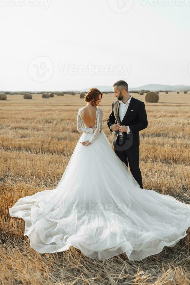The bride and groom are standing in the field, and behind them are large sheaves of hay. The bride stands with her shoulders turned to the camera. Long elegant dress. Stylish groom. Summer photo