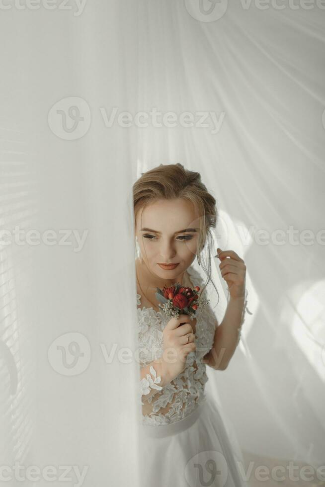 Preparation for the wedding. Beautiful young bride in white wedding dress indoors. Luxury model looks at wedding shoes, at home in studio room with big window. The girl shows photo