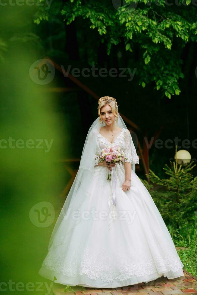 amplio retrato de el novia. el novia es en pie en un blanco vestir y velo con un ramo de flores en contra el antecedentes de lozano verde arboles un maravilloso vestido. bonito mujer. Boda foto