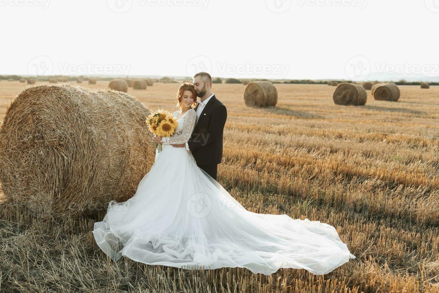 Wedding portrait of the bride and groom. The groom hugs the bride from behind, next to a bale of hay. Red-haired bride in a long dress with a bouquet of sunflowers. Stylish groom. Summer photo