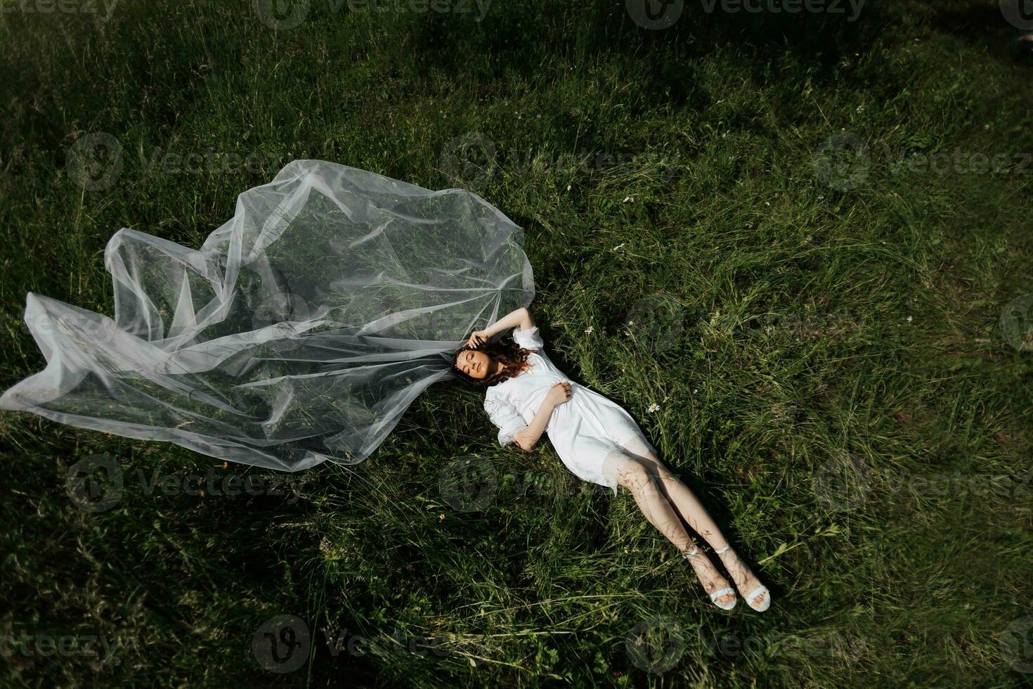 Morning of the bride in nature. The bride is in a dressing gown and a long veil, lying on the green grass and enjoying the sun's warmth. Wide angle photo. photo