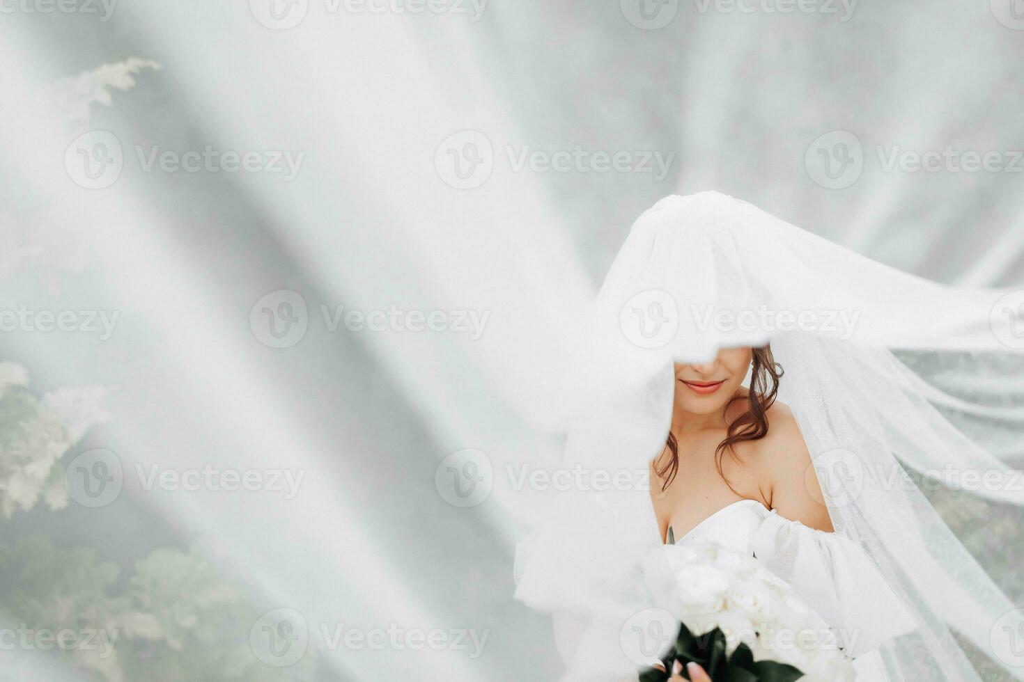 Curly brunette bride in a white dress, covered with a veil, poses for the camera with a bouquet of roses. Portrait of the bride. Beautiful makeup and hair. Wedding in nature photo