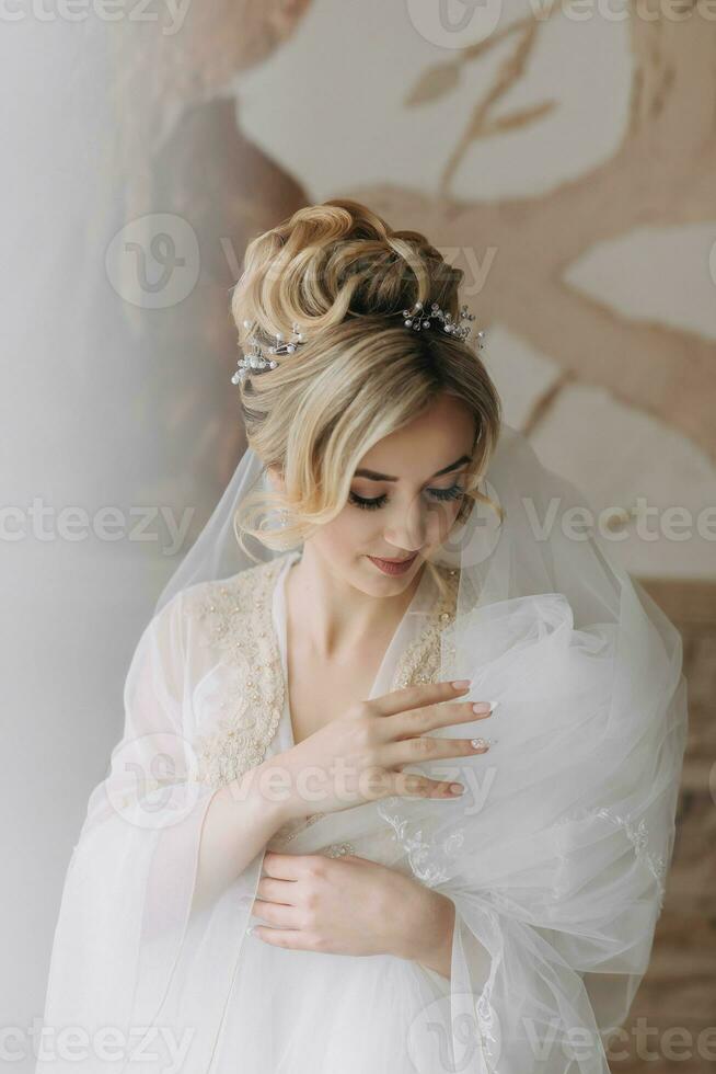A bride with an elegant hairstyle, in a robe, stands looking down and holding her snow-white veil against the background of painted walls. photo
