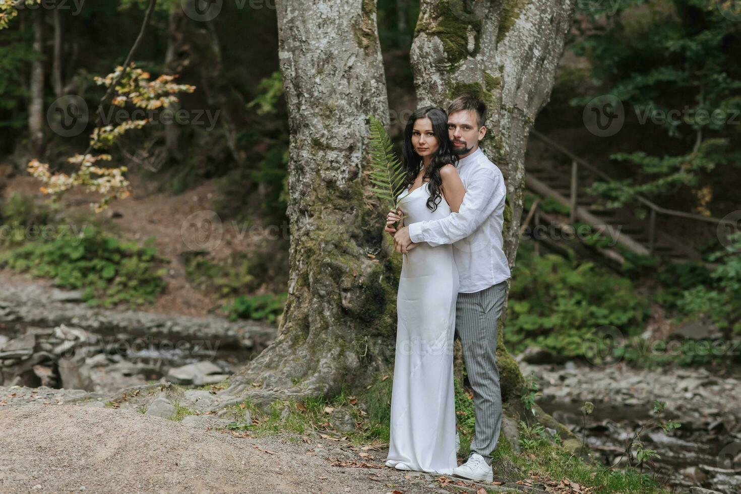 A girl with long black curly hair, standing in a white silk dress in a spring forest against a large tree, holds a fern branch in her hands. Behind her is her husband photo