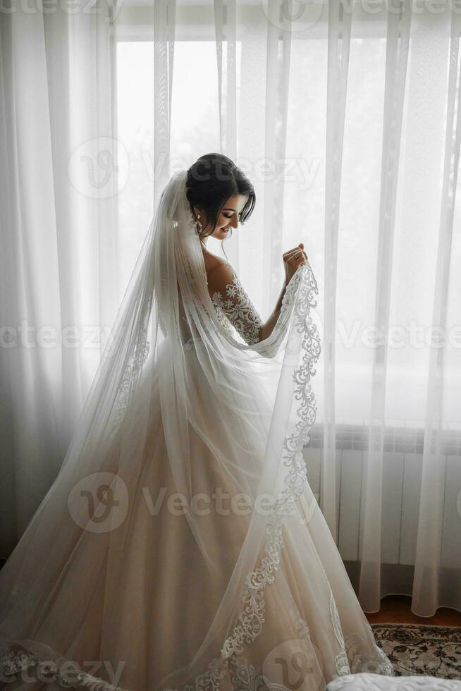 A brunette bride in a white dress and a long veil poses standing by the window. Beautiful hands. French manicure. Beautiful hair and makeup. Daylight photo