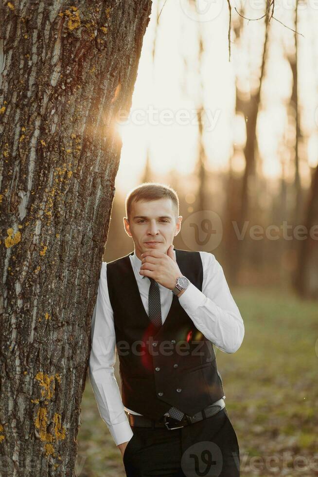 Portrait of the groom. The groom in a black vest is standing in the forest, leaning on a tree and touching his chin, looking into the camera lens. Wedding in nature photo