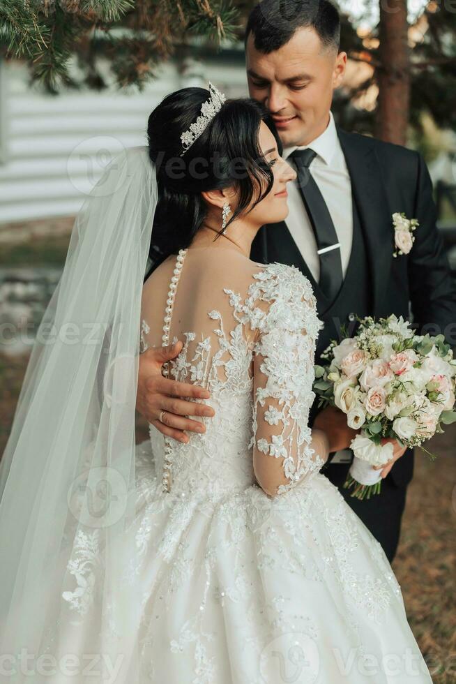 Wedding portrait in nature. The brunette bride and groom in a white long dress are standing and hugging against the background of coniferous trees and a white hut. Stylish groom. photo