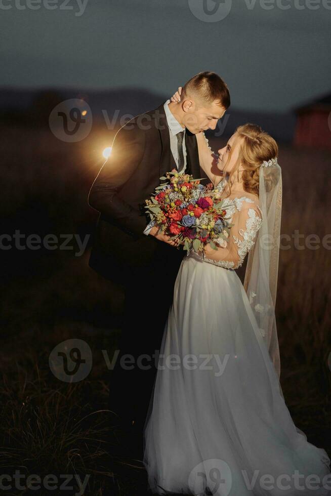 Beautiful bride and groom in evening lights, kissing and hugging holding a bouquet of flowers photo